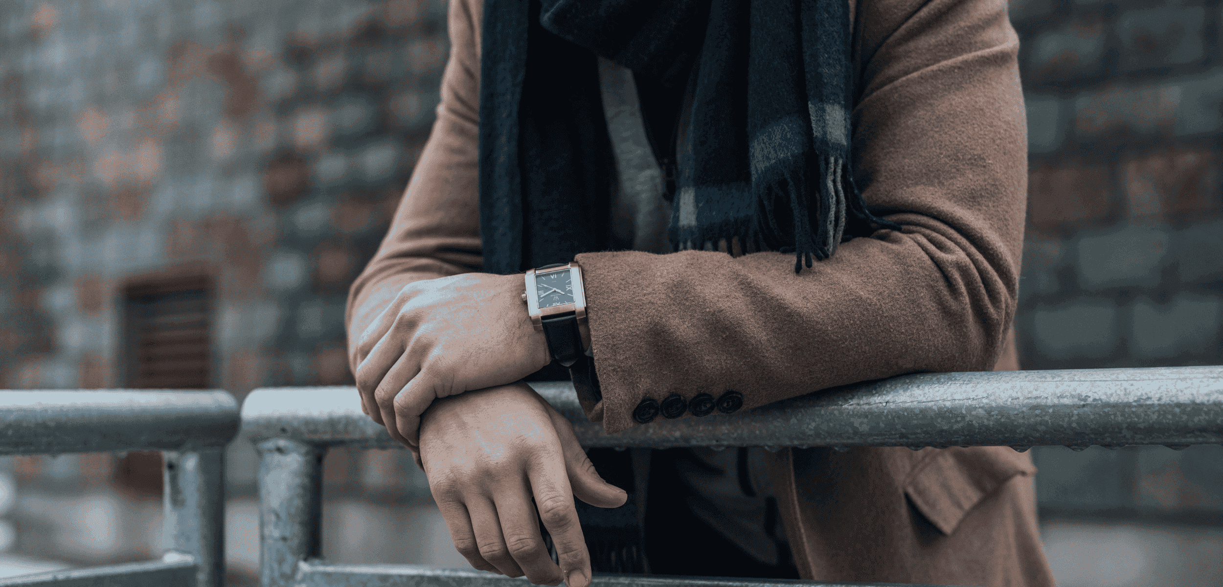 A person wearing a brown coat and a blue scarf leans on a metal railing. They have a wooden watch with a blue face on their wrist. The background features a brick wall.