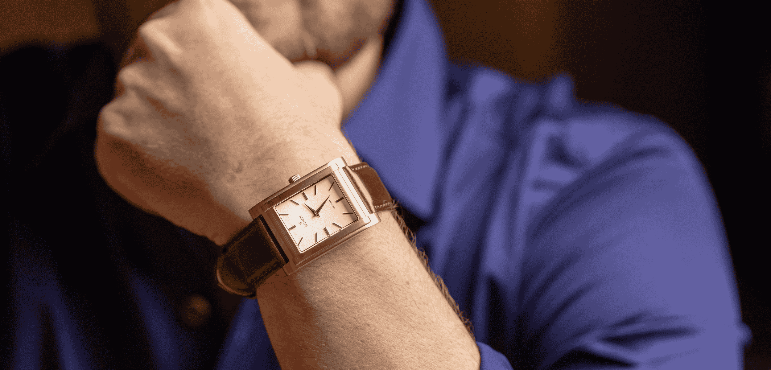 A person wearing a blue shirt, resting their chin on their hand, is showcasing a rectangular watch with a leather strap. The watch features a minimalist white dial adorned with thin black hour markers and hands.