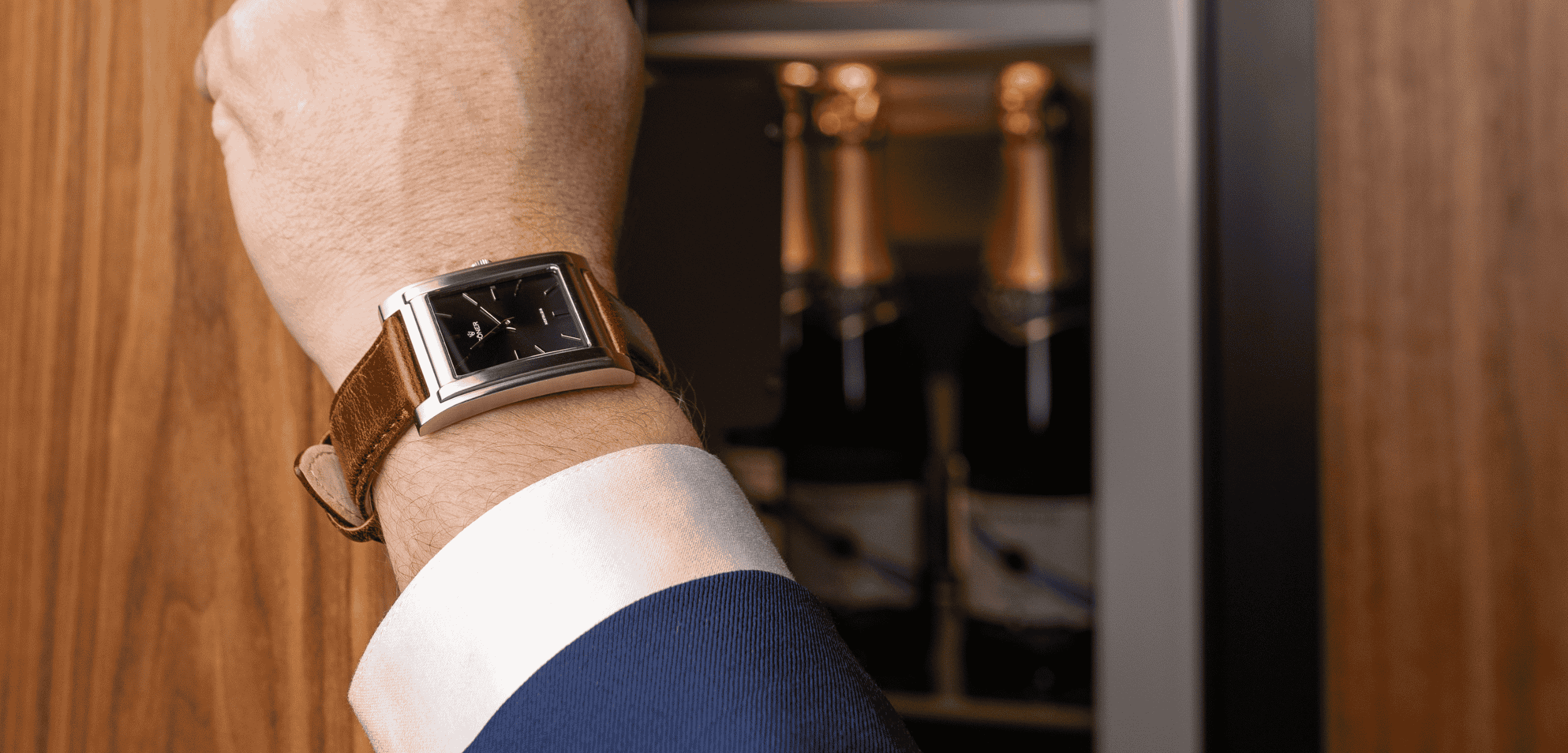 A person wearing a rectangular watch with a brown leather strap reaches towards a wooden cabinet. Inside the cabinet, several bottles of champagne are visible.