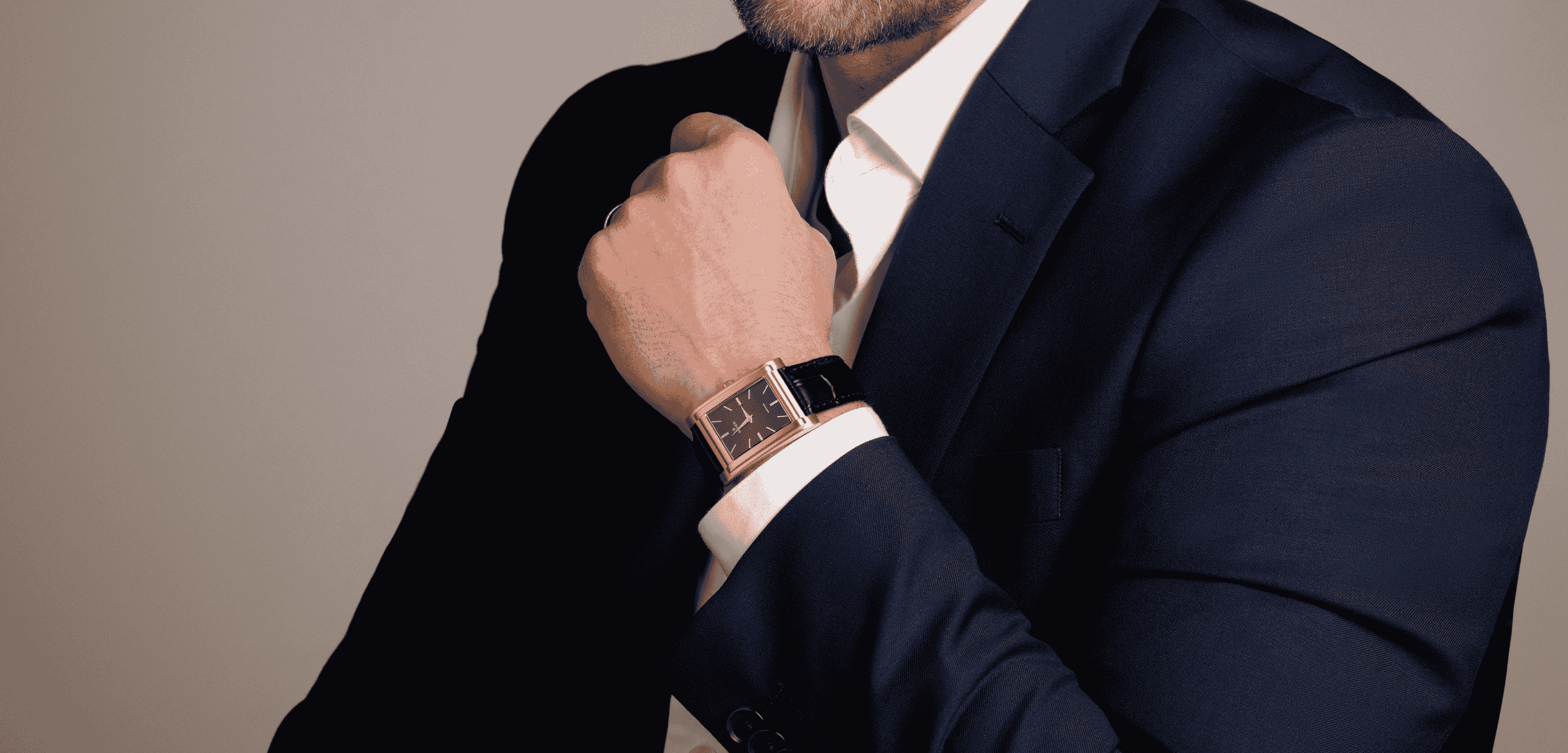 A person wearing a dark blue suit and white shirt displays a wristwatch with a gold rectangular face and black strap. The persons face is not visible.