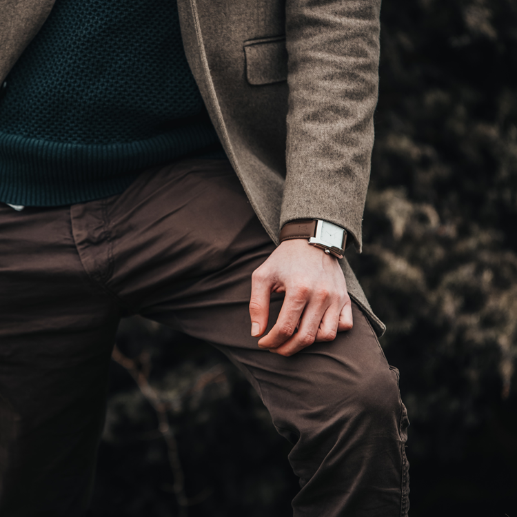 A man wearing a brown blazer, teal sweater, and brown pants complements his look with a stylish rectangular watch. The background is blurred with greenery, enhancing his sophisticated appearance.