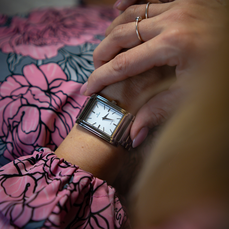 Close-up of a person wearing a silver rectangular wristwatch with a white dial, resting their hand on another hand. The person is wearing a pink and black floral-patterned blouse, and a thin silver ring is visible on one finger.