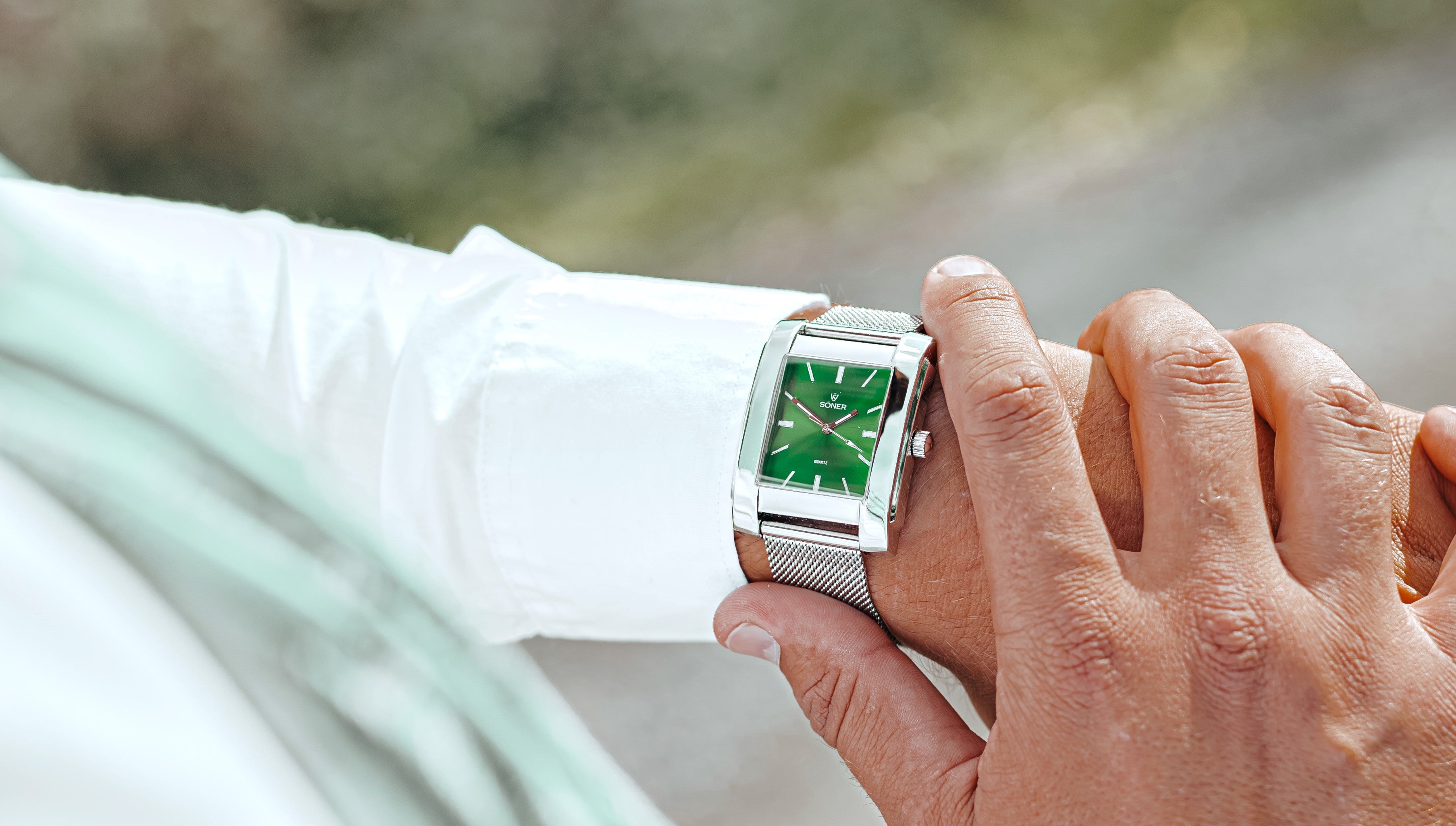 A person wearing a silver mesh band watch with a green rectangular face, checking the time on their wrist. The person is dressed in a white shirt, and the background is softly blurred, suggesting an outdoor setting.