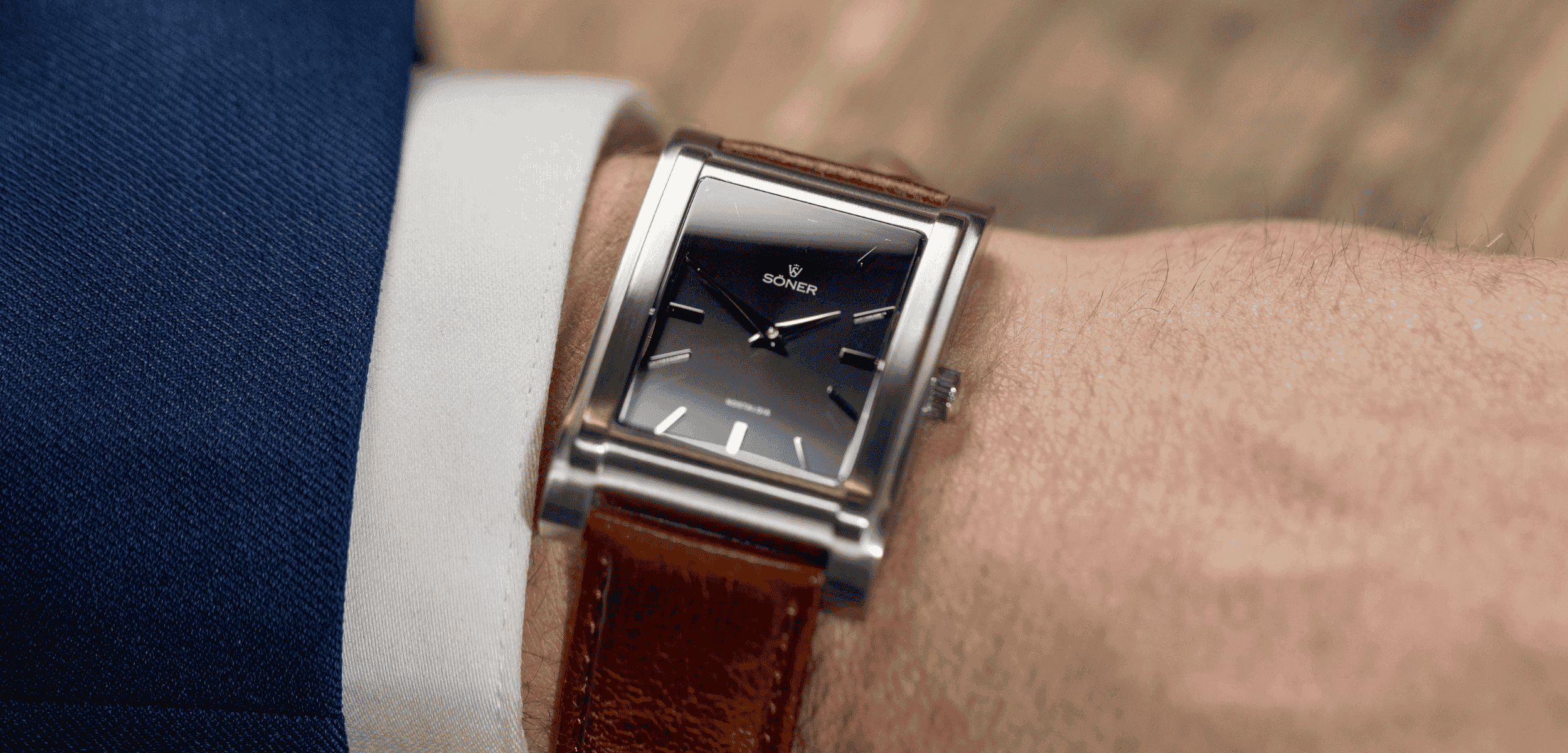 Close-up of a persons wrist wearing a square silver watch with a black face and brown leather strap. The person is dressed in a blue suit jacket and white shirt. The background is a blurred wooden surface.