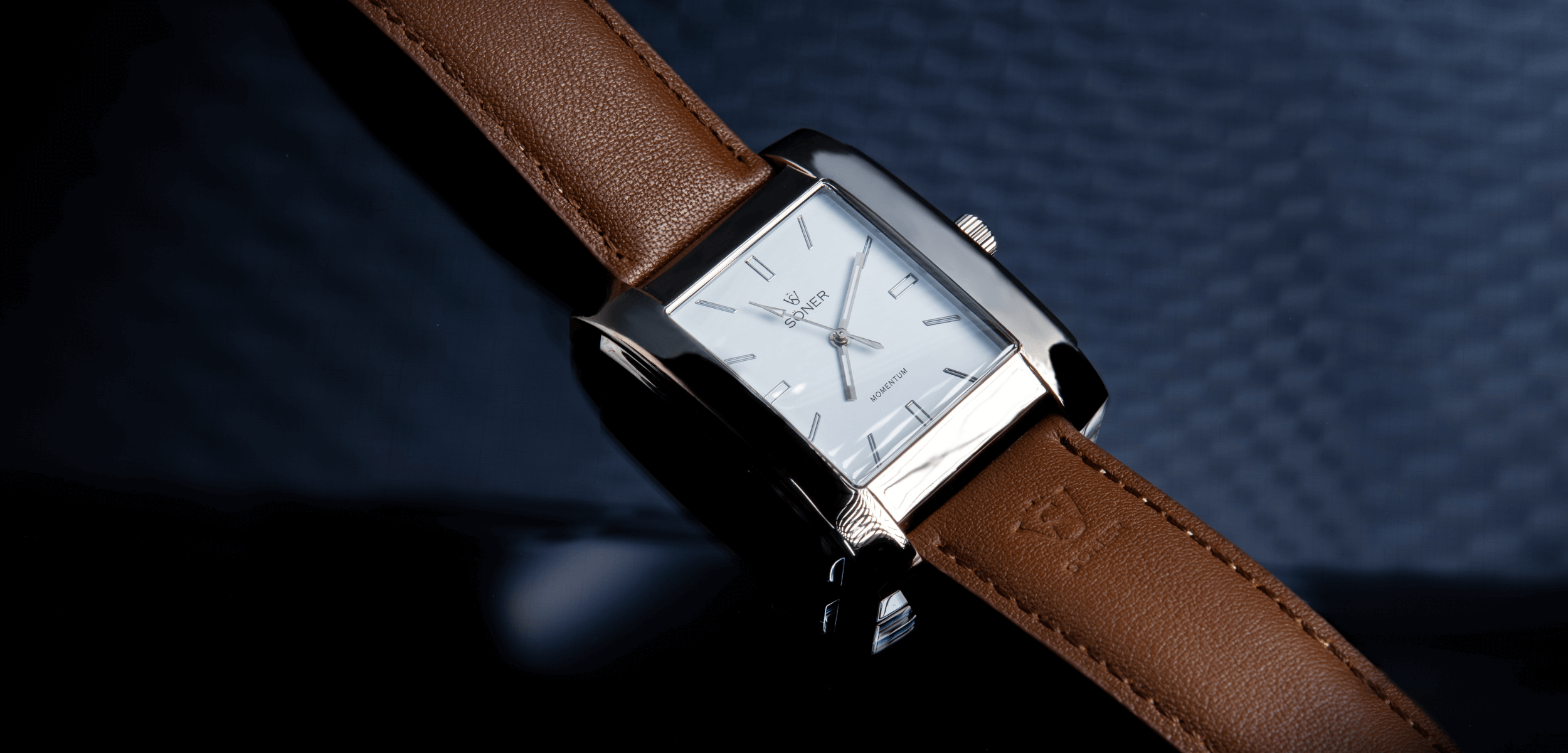 This rectangular silver watch boasts a white dial with minimalist hour markers and hands. Complemented by a brown leather strap, it stands out against a dark, textured background.