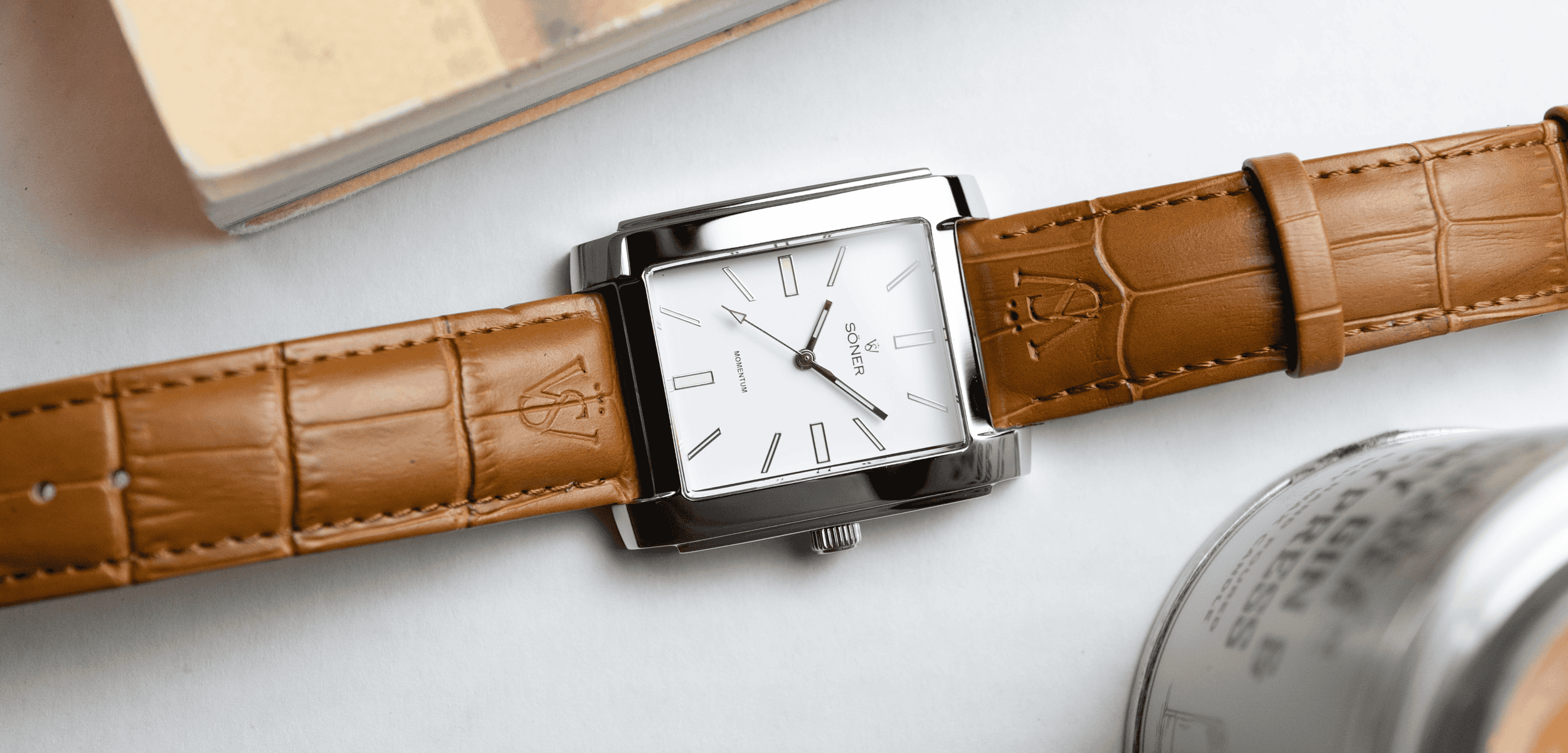 A rectangular watch with a white face, silver casing, and brown leather strap is placed on a white surface. The watch has simple hour markers and three hands, and is positioned horizontally. Nearby are a book and a metallic container.