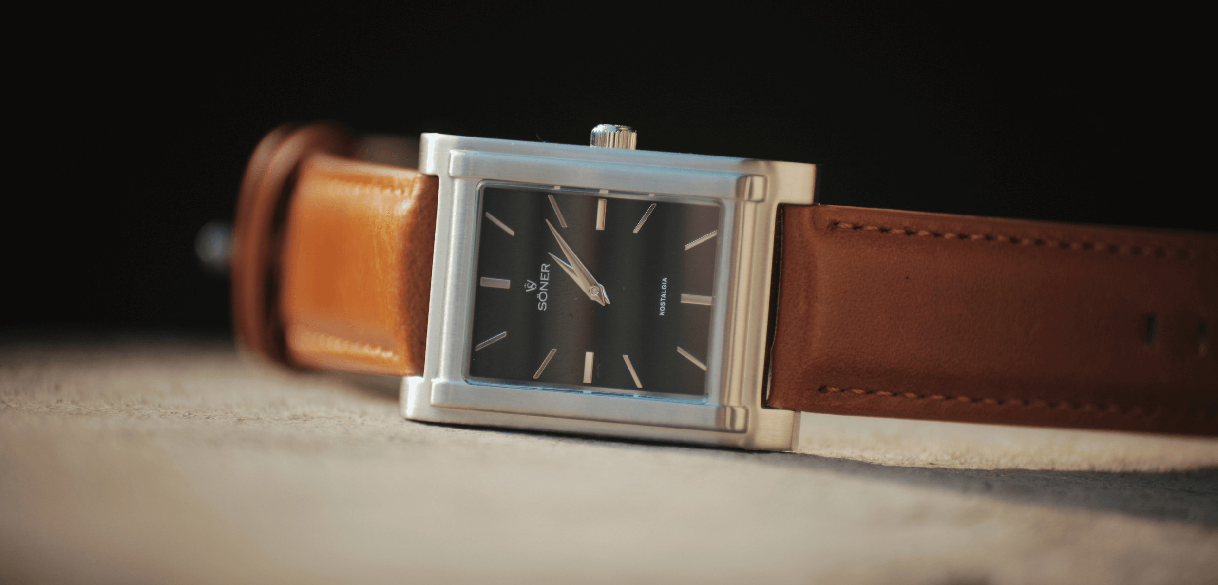 A rectangular watch with a brown leather strap rests on a textured beige surface. Its black face, adorned with silver hour markers and hands, is enclosed in a metallic frame. The dark background creates striking contrast, highlighting the elegance of this timepiece.