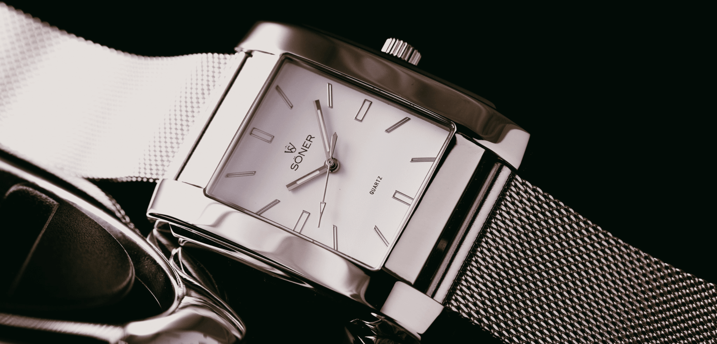 A sleek, rectangular silver wristwatch with a mesh strap sits against a dark background. This rectangular watch features a minimalist face with silver hour, minute, and second hands and a simple index, marked with QUARTZ and a logo.